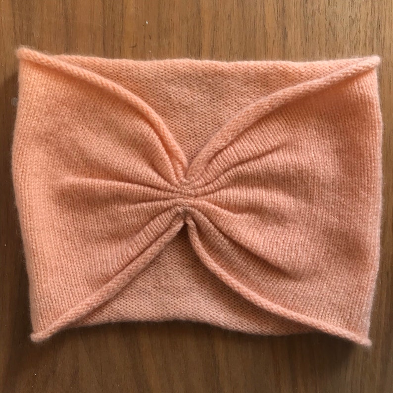 May include: A peach-colored knitted headband with a gathered center detail. The headband is made of a soft, stretchy fabric and is designed to fit comfortably around the head.