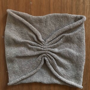 May include: A gray knitted headband with a gathered center and a folded design. The headband is made of a soft, textured fabric.