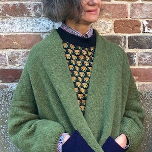 May include: An olive green knit cardigan featuring long sleeves with dark navy blue ribbed cuffs, an open front design, and a relaxed fit. The garment is layered over a black shirt with a repeating yellow floral pattern and a patterned collar.