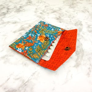 May include: A teal fabric pouch with a floral pattern and a red fabric flap with a geometric pattern. The pouch is open and a white pill pack is inside.