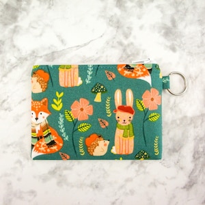 May include: A teal zippered pouch with a whimsical woodland print featuring foxes, rabbits, and hedgehogs wearing scarves and hats. The print also includes flowers, leaves, and mushrooms.