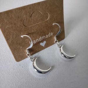 May include: Silver crescent moon earrings with a curved hook design. The earrings are presented on a brown card with the word "handmade" and a heart symbol. The background is a light, neutral colour.