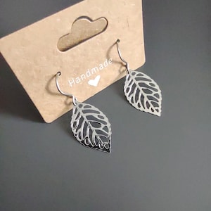 May include: Silver leaf earrings with a delicate, openwork design. The earrings are displayed on a brown card with the word "Handmade" and a heart symbol. The earrings have a hook closure.
