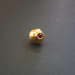 May include: A small, spherical gold bead with a textured, hammered surface finish. The bead features a single, circular, faceted red gemstone set securely into the centre of its rounded exterior, creating a distinct contrast against the gold.