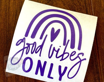 May include: A white sticker with a purple rainbow design and the words "good vibes ONLY" in a handwritten font. A small purple heart is above the word "ONLY". The sticker is on a wooden surface.