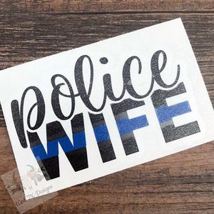 Puede incluir: Adhesivo blanco con las palabras "police WIFE" en escritura negra y letras de bloque. La palabra "WIFE" tiene un diseño de rayas azules. El adhesivo está sobre una superficie de madera.