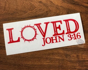 May include: A white rectangular sticker with red text. The word "LOVED" is at the top, with a crown of thorns in the center. Below is "JOHN 3:16". The sticker has a glossy finish.