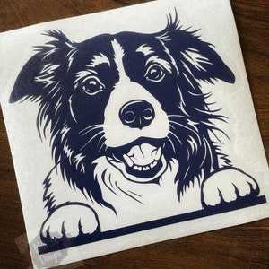 May include: A black and white border collie dog with a blue outline peeks over a white surface. The dog has a happy expression with its tongue sticking out.