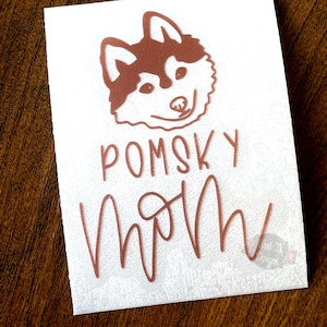 May include: A white sticker with a copper-colored husky dog illustration and the text "Pomsky Mom" in a cursive font.