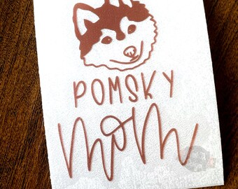 Könnte beinhalten: Ein weißer Aufkleber mit einer kupferfarbenen Husky-Hund-Illustration und dem Text "Pomsky Mom" in einer kursivschrift.