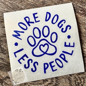 Puede incluir: Una calcomanía de vinilo blanca cuadrada con texto azul oscuro que dice MORE DOGS LESS PEOPLE dispuesto en un círculo alrededor de un icono de huella con una forma de corazón en el centro. Un pequeño logotipo cuadrado con el texto West TX Designs es visible.