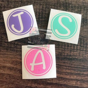 May include: Three circular vinyl decals with white outlines. Each decal features a different letter: a purple "J", a teal "S", and a pink "A". The decals are on a wooden surface. The text "WEST TX DESIGNS" is partially visible.