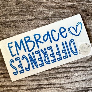 May include: A white rectangular decal featuring the blue text EMBRACE and the word DIFFERENCES printed in a mirrored orientation below it. A blue heart outline is positioned to the right of the word EMBRACE. A small circular logo reads West TX Designs.