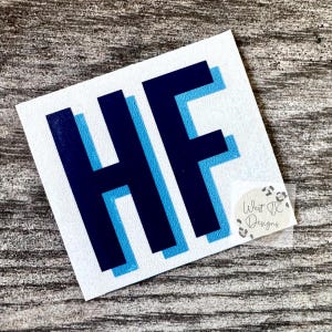 May include: A square white decal featuring the letters HF in a layered design. The letters are rendered in a dark navy blue color with a light blue shadow effect behind them. A small circular sticker with the text West TX Designs is attached.