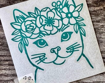 May include: A teal vinyl decal featuring a line art illustration of a cat wearing a floral crown of roses and leaves. The cat has large eyes and prominent whiskers. A small square sticker with the text West TX Designs is in the bottom corner.