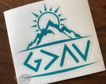 May include: A teal vinyl decal on a white backing featuring a stylized mountain range beneath a sun with radiating rays. Below the mountains, the text G>AV is displayed in a bold, sans-serif font, underlined by a horizontal line.
