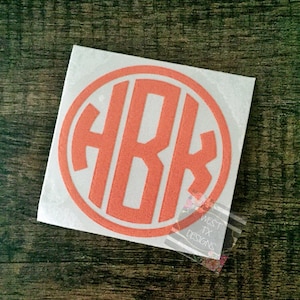 May include: A square white sticker with an orange monogram inside an orange circle. The monogram features the letters H, B, and K. The sticker is on a dark wood surface. The text "WEST TX DESIGNS" is visible on a clear sticker in the corner.