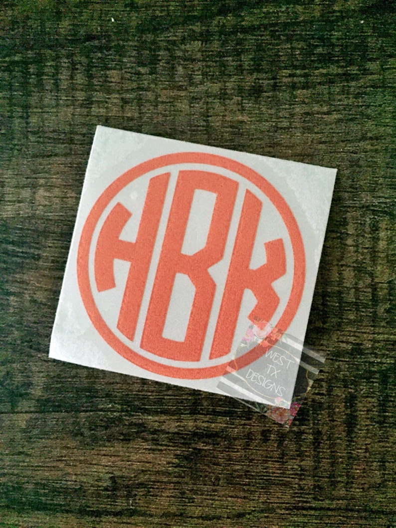 May include: A square white sticker with an orange monogram inside an orange circle. The monogram features the letters H, B, and K. The sticker is on a dark wood surface. The text "WEST TX DESIGNS" is visible on a clear sticker in the corner.