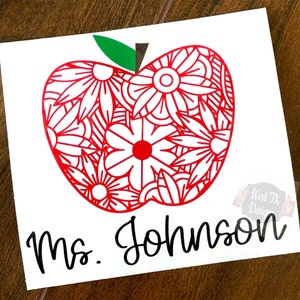 Puede incluir: Una manzana roja con un diseño floral blanco y un tallo verde. El texto "Ms. Johnson" está escrito en negro debajo de la manzana.