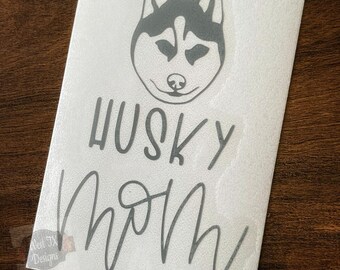 May include: A white sticker with a gray husky dog face and the words "HUSKY MOM" in a handwritten font. The sticker is on a dark brown wooden surface.