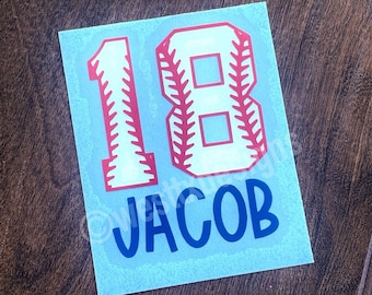 May include: A blue and white baseball jersey with the number 18 in red and white. The name "JACOB" is printed in blue below the number.