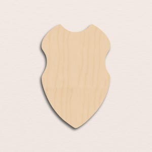 Puede incluir: Un escudo de madera de color claro con una parte superior curva y una parte inferior puntiaguda. La veta de la madera es visible y el escudo no está pintado. El escudo es un lienzo en blanco para manualidades o decoración.