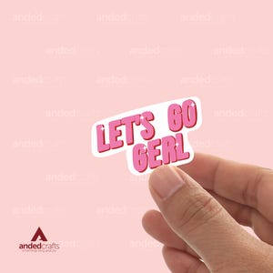 May include: A pink and white sticker with the text "LET'S GO GERL" in a bold, bubble-style font. The sticker is held by a hand against a light pink background. The sticker is a fun, motivational item.