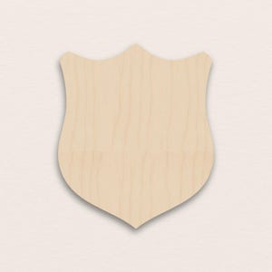 May include: A wooden shield-shaped cutout with a light tan color. The shield has a curved top and a pointed bottom, with a smooth surface and visible wood grain. The cutout is designed for crafting or decorative purposes.