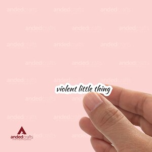 Può includere: Un adesivo bianco con testo nero che dice "violent little thing" su uno sfondo rosa.