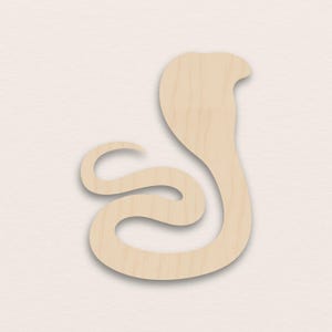 Puede incluir: Un recorte de madera de una cobra en color beige claro. La serpiente se representa en una pose estilizada y curvada, con la cabeza levantada y la capucha ligeramente abierta. La veta de la madera es visible, agregando textura al diseño.
