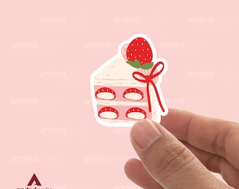 Könnte beinhalten: Ein Aufkleber mit einem rosa und weißen Kuchen mit einer Erdbeere oben drauf. Der Kuchen ist mit einem roten Band verziert. Der Aufkleber wird von einer Hand gehalten.