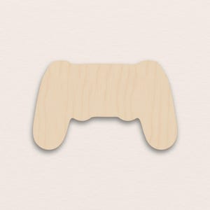Puede incluir: Recorte de madera de un mando de videojuegos. El mando es de color marrón claro con una superficie lisa y bordes redondeados. El diseño presenta dos asas y un cuerpo central con una base curva.