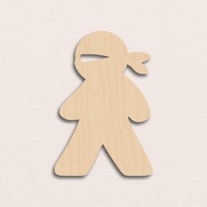 Puede incluir: Recorte de madera de una figura de ninja, con una cubierta para la cabeza con una ranura para los ojos y una cola de caballo. La figura tiene los brazos y las piernas extendidos y está hecha de madera clara.