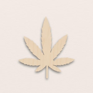 Op de afbeelding: Een houten uitgesneden cannabisblad. Het blad is lichtbruin met een natuurlijke houtnerf. Het heeft zeven punten en een steel. De achtergrond is effen, gebroken wit.