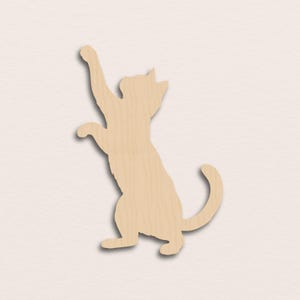 Op de afbeelding: Een platte, lichtgekleurde houten uitsparing in de vorm van een kat die op de achterpoten staat met één voorpoot omhoog. Het silhouet heeft een gladde, natuurlijke houtnerftextuur en een gekrulde staart die naar de rechterkant uitsteekt.