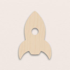 May include: A wooden rocket ship cutout with a circular hole near the top. The rocket has a light wood grain finish and two curved fins at the bottom. The cutout is a light tan color.