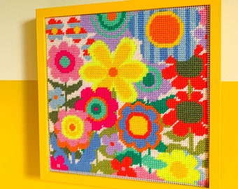 May include: A framed cross-stitch artwork featuring a colourful floral design with yellow, red, blue, green, and pink flowers. The artwork is framed in a yellow wood frame.