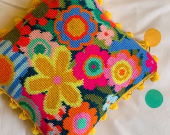 May include: A colourful floral patterned cushion with a yellow pom-pom trim. The cushion is made of a soft, plush fabric and features a variety of bright colours, including pink, orange, yellow, green, and blue.