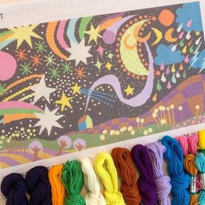 Könnte beinhalten: Eine Gobelin-Leinwand mit einem farbenfrohen Himmelsdesign, das Sterne, Regenbogen und einen Mond zeigt. Das Wort "STARDUST" ist links oben aufgedruckt. Darunter befinden sich Bündel farbigen Stickgarns in verschiedenen Farbtönen.