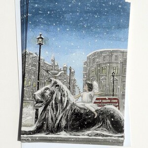 May include: A snowy winter scene with a white angel riding a black lion statue. The angel is wearing a white robe and has wings. The lion is sitting on a snow-covered street with a red double-decker bus in the background. The scene is lit by a street lamp.