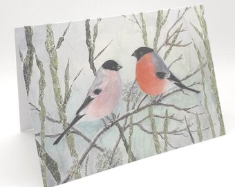 May include: A greeting card featuring a watercolor illustration of two birds perched on branches in a snowy forest. The birds are a pink and gray bird and a red and gray bird.