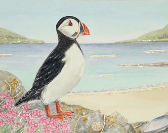 May include: Watercolor painting of a puffin bird perched on a rock, overlooking a beach and ocean. The bird has black and white feathers, a red beak and orange feet. Pink flowers and gold accents are in the foreground.