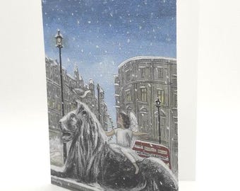 May include: A watercolor illustration of a snow scene in London, England. A white angel figure sits on a stone lion statue in front of a red double-decker bus. The scene is set against a backdrop of snow-covered buildings and a blue sky with falling snow.