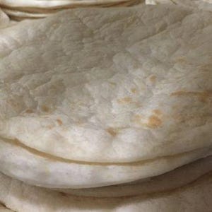 May include: A stack of fresh, round tortillas. The tortillas are a light beige color with slightly browned spots, and have a slightly textured surface. The stack is on a metal rack, with more tortillas visible in the background.