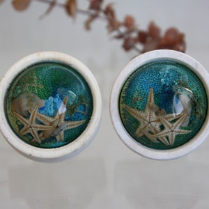 Può includere: Un paio di orecchini in ceramica bianca con una cupola in resina trasparente che contiene uno sfondo blu e verde con due stelle marine.