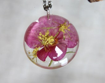 May include: A clear, spherical pendant necklace with a pink flower and yellow details set in resin. The pendant is suspended from a silver-coloured chain. The necklace is shown against a blurred backdrop.