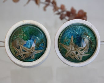 May include: A pair of white ceramic earrings with a clear resin dome containing a blue and green background with two starfish.