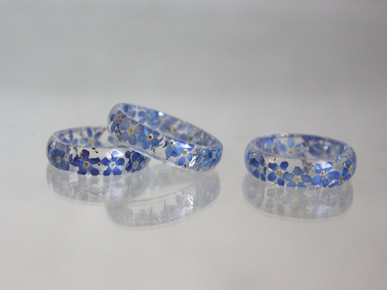 May include: Three clear resin rings with embedded blue forget-me-not flowers. The rings are displayed on a reflective surface, showcasing the delicate floral design. The rings are a unique accessory.