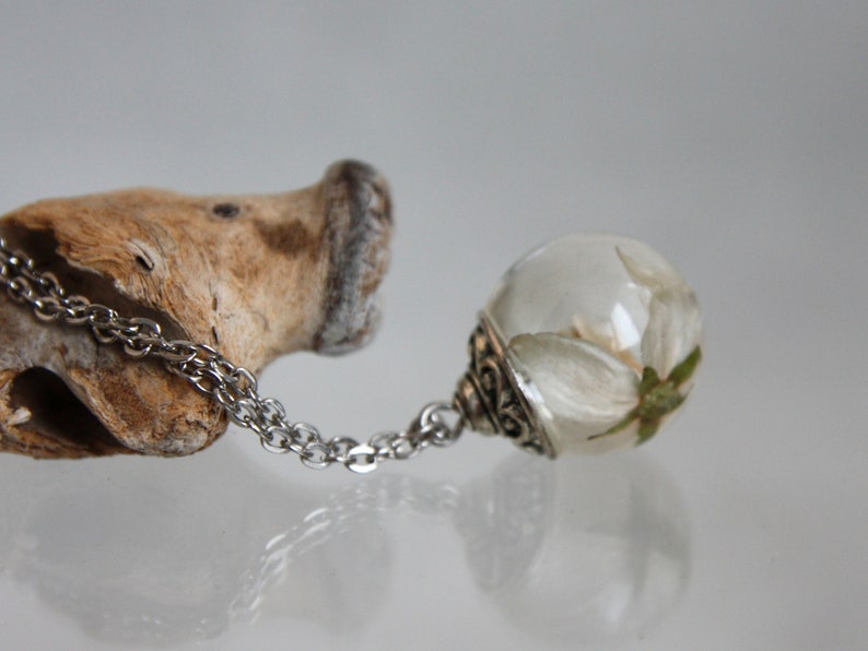 May include: A silver chain necklace with a clear glass pendant containing a white flower. The pendant has a silver ornate design around the edge.