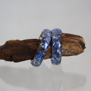 May include: Two clear resin bangles with blue forget-me-not flowers and silver flakes embedded inside. The bangles are displayed on a piece of driftwood against a white background. The floral design adds a touch of nature-inspired elegance.